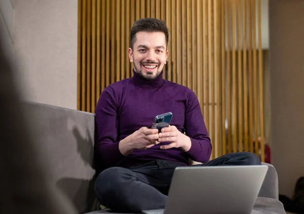 Усміхнений чоловік з ноутбуком на дивані говорить на мобільному телефоні . — стокове фото