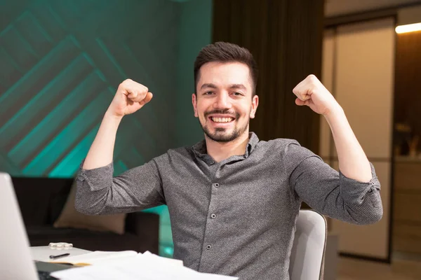 El hombre sonriente levantó alegremente las manos. Trabajar desde casa sentado en una mesa con un portátil y documentos. — Foto de Stock