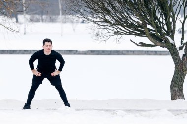 Genç adam sabah egzersizlerinde esneme hareketleri yapıyor, kışın dışarıda, karda spor yapıyor..