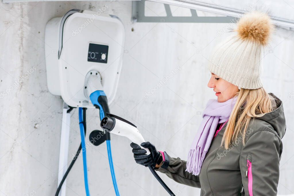 charging station, ecological fuel concept. Portrait of a beautiful young girl holding a charging plug in her modern electric car for refueling at a city station.