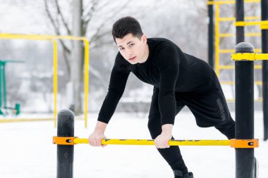 Genç adam sabah egzersizleri yapıyor, kışın dışarıda, karda, sarı egzersiz makinesinin yardımıyla spor yapıyor..