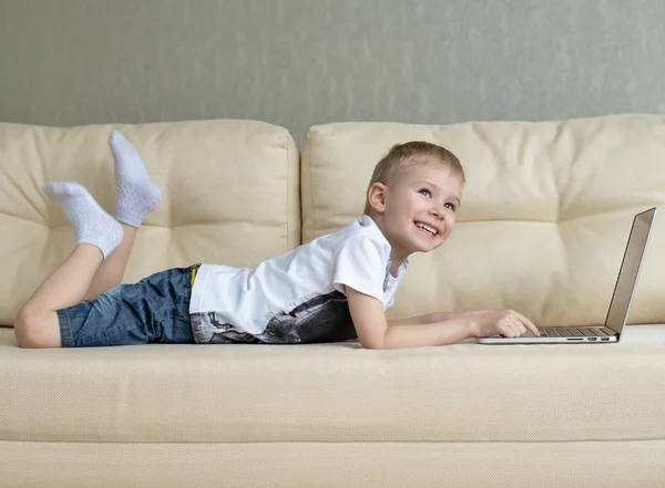 Retrato de un niño pequeño estirado en el sofá delante de la computadora portátil —  Fotos de Stock