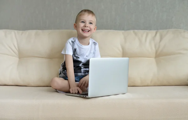 Niño pequeño sentado con el ordenador portátil en el sofá en casa —  Fotos de Stock