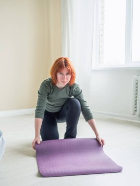 Kız pencerenin yanındaki paspası kaldırıyor ve kameraya bakıyor. Yoga ve egzersiz yapmaya hazırlanan bir kadın. Ön görünüm.