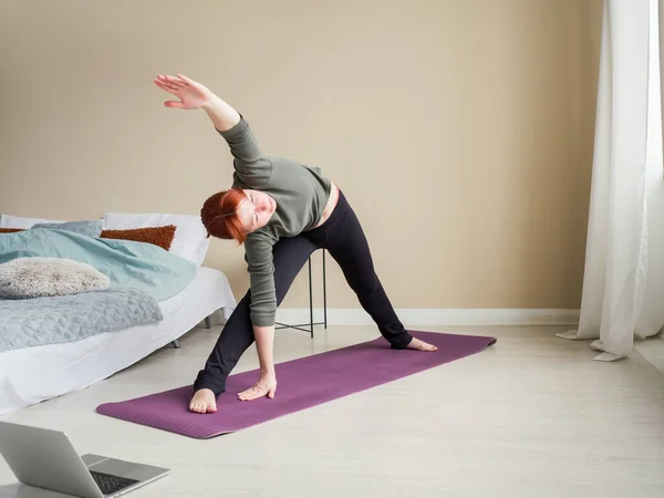 Treinador de cabelo vermelho menina esticando no tapete de exercícios perto da cama. Treinamento de vídeo online usando laptop. Conceito de estilo de vida saudável. Vida ativa em auto-isolamento. — Fotografia de Stock