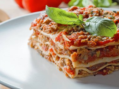 Extra close up view to piece of lasagna. Dish served basil leafs. Tomatoes on backdrop. Menu of Italian cuisine. Side view.