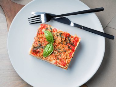 Piece of lasagna. White plate with fork and knife. Basil leafs, tomatoes and zucchini on table. Menu of Italian cuisine.