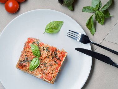 Top down view to piece of lasagna. Basil leafs, tomatoes and zucchini on table. Menu of Italian cuisine.