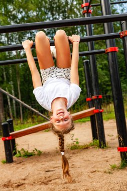 Spor üniformalı bir kız turnikede baş aşağı asılı duruyor. Spor sahası, hücum, ısınma, gündüz, açık hava.