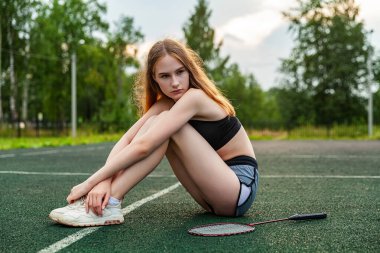 Spor tişörtlü ve şortlu genç ve güzel bir kız spor sahasında oturuyor. Gündüz vakti, fitness, açık hava, badminton, raket