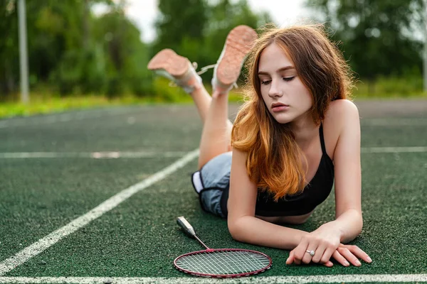 Young beautiful girl in sports top and shorts lying on the sports ground, posing. Daytime, fitness, open air, badminton, racket