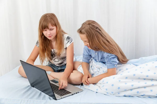 Two girls girlfriends with long blond hair in home clothes play on a laptop, sitting on the bed. Distance learning. Lessons. homework. education. laptop