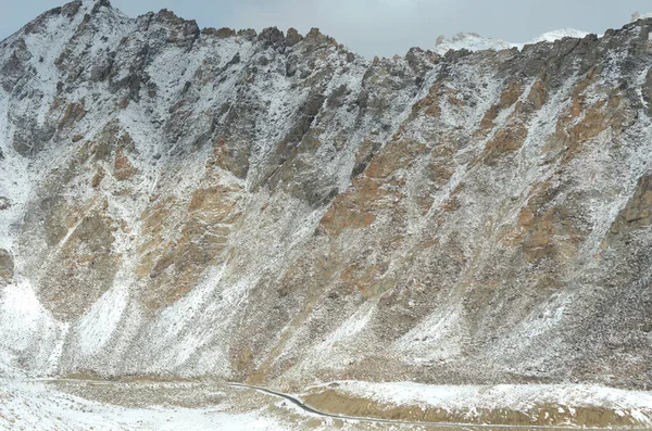 在印度 一条路沿着积雪覆盖的山脉的底部走着 汽车是看得见的 群山的山峰升到阴沉沉的天空中 — 图库照片