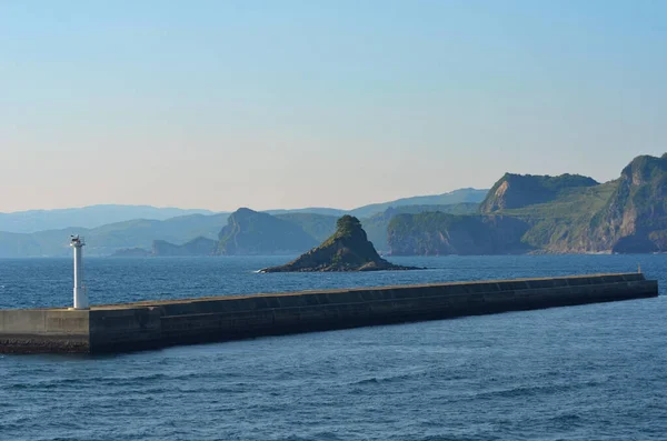 Bir deniz feneri taştan bir duvarın üzerinde, suyla çevrili. Kıyıyla tepe arasında küçük kayalık bir çıkıntı var. Gökyüzü mavi, pembe dokunuşlarla.