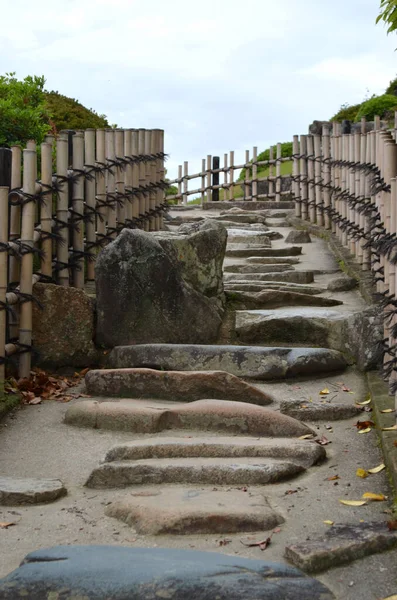 A path lines with stones leads over a hill. A bamboo fence separates the path from grass and bushes. The sky is overcast.