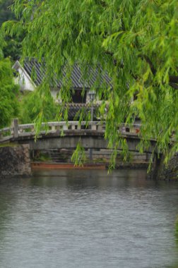 Taştan bir köprü zarif bir şekilde nehrin üzerinde kıvrılır. Ön planda, bir söğüt ağacı manzarayı çerçeveler. Arka planda gri çatılı bir ev ve küçük ahşap bir tekne var..