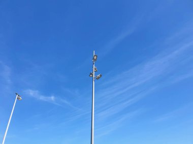 Sokak lambalı uzun metal direkler aşağıdan görülüyor. Bir direkte dört ışık var, diğerinde. Gökyüzü soluk beyaz bulutlarla mavidir..