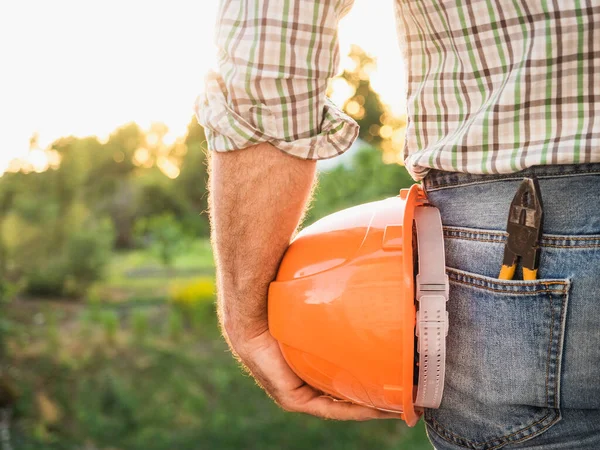 Attractive man in work clothes, holding tools in his hands against the background of trees, blue sky and sunset. View from the back. Labor and employment concept