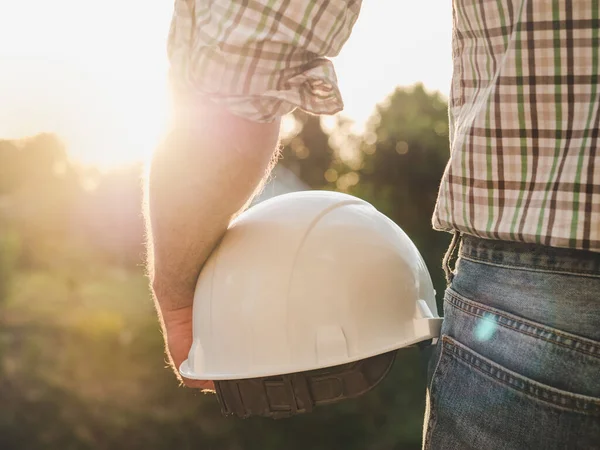 Attractive man in work clothes, holding tools in his hands against the background of trees, blue sky and sunset. View from the back. Labor and employment concept