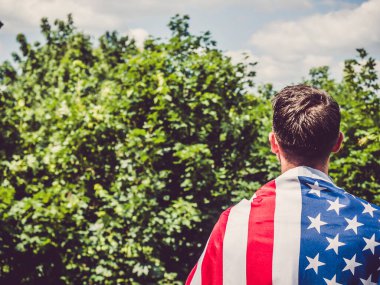 Ağaçların, mavi gökyüzünün ve günbatımının arka planında Amerikan bayrağını tutan yakışıklı bir adam. Arkadan bak. Çalışma ve istihdam kavramı