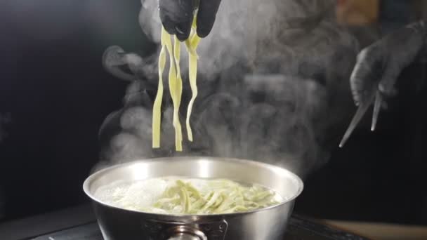 Chef in black gloves putting raw pasta into boiling water. White smoke in slow motion rising above hot saucepan on black background. Cooking in restaurant. Delicious Italian pasta. Full hd — Stock Video