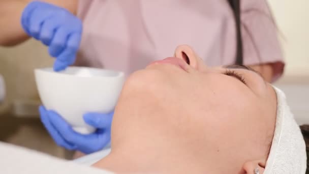 Conceito de clínica de beleza. Médica cosmetologista feminina preparando máscara facial para o procedimento de mistura em tigela usando escova perto do paciente. agitando componentes em tigela de vidro. 4 k vídeo — Vídeo de Stock