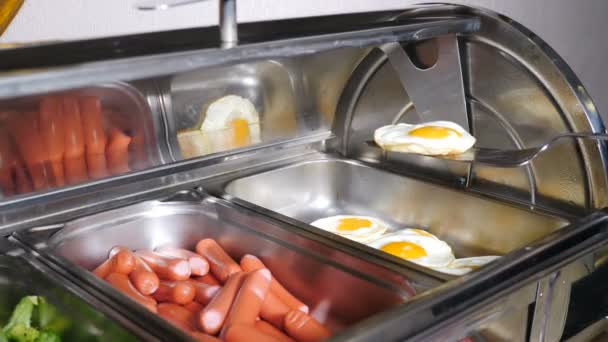 Podgrzewane tacki z kiełbaskami, gotowane brokuły i smażone jajka w hotelowej luksusowej restauracji. Włącznie. Bufetowe jedzenie. Kelner umieszcza kawałek smażonego jajka. Hotel zaprasza na śniadanie w formie bufetu. 4 tys. wideo — Wideo stockowe
