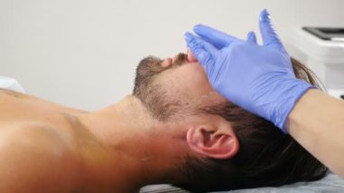 Refah, sağlık hizmeti konsepti. Güzellik kliniğindeki kanepede yatan adam yüz bakımı yaptırıyor. Yüz temizleme prosedüründe yakışıklı bir erkek müşteri. Cilt bakımı. 4 k video