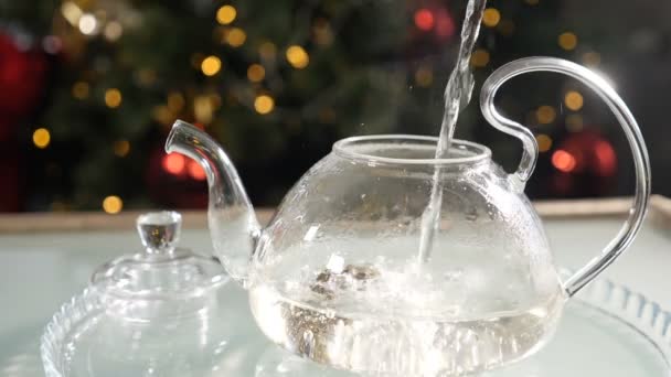 Tiempo de Navidad y ambiente de Año Nuevo. Preparación de té con agua hirviendo con abeto de Año Nuevo en el fondo. Haciendo té en cámara lenta. Añadir agua a la flor de té de jazmín en la tetera de vidrio. Full hd — Vídeos de Stock