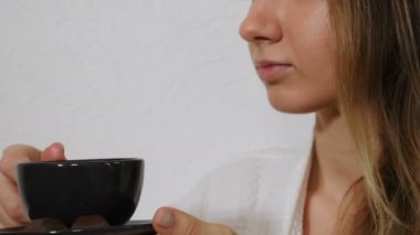 Beyaz bornozlu genç bir kadın spa ameliyatından sonra çay, kahve ya da çikolata içiyor. Kadın eli kupayı ağzına götürür ve içecek içer. Sabah çayı içiyorum. 4k video