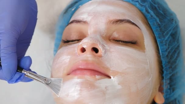 Klinika urody opieki zdrowotnej. Młoda atrakcyjna kobieta otrzymuje profesjonalną pielęgnację twarzy. Kosmetyk nakłada maskę na twarz pędzlem. Kosmetolog rozprowadza krem na kobiecej twarzy przed zabiegiem — Wideo stockowe