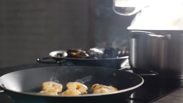 Cooking in restaurant kitchen. Prawns being cooked in frying pan with steaming saucepan and pan in background. Steam rising up in slow motion. chef pouring broth from ladle into seafood. Full hd — Stock Video