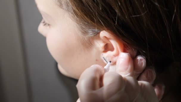 Jolie adolescente se faisant percer les oreilles. Cosmetologue préparant les oreilles du patient pour le processus de perçage. Créer des zones pour les futures boucles d'oreilles. Travailleur médical faisant un trou dans le cartilage de l'oreille. Full hd — Video