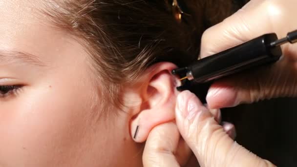 En söt tonårstjej med hål i öronen. Kvinnlig patient som får öron genomborrade av kosmetolog. Medicinsk arbetare gör hål och placera örhänge i brosk i örat med enhet. Fullständig hd — Stockvideo