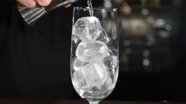 Barman maakt cocktail gieten transparante alcohol vloeistof in glas met ijsblokjes. Langzame beweging. Sluit maar af. Zwarte achtergrond. Er stroomt nog steeds water. Spatten en druppels. Vol hd — Stockvideo