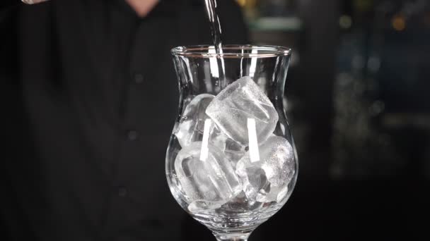 Cantinero haciendo cóctel verter líquido de alcohol transparente en vidrio con cubitos de hielo. En cámara lenta. De cerca. Fondo negro. Todavía está lloviendo agua. salpicaduras y gotas. Full hd — Vídeo de stock