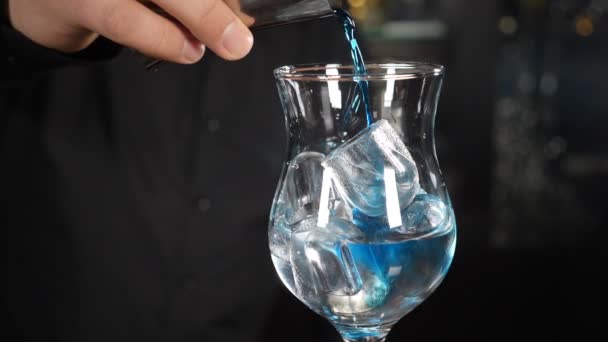 青いカクテルだ。バーカウンターで爽やかなカラフルなカクテルを作るバーテンダー。バーマンは黒いテーブルの上に氷でいっぱいのガラスに青い夏のカクテルを注ぐ。アルコールカクテルパーティーナイトライフ — ストック動画