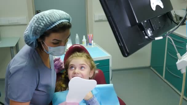 Petite jolie fille et dentiste féminine regardant miroir après un traitement des dents réussi et riant. Des médecins amicaux. Bureau de dentiste moderne. Enfant sur chaise dentaire. Vidéo 4 k — Video