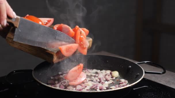 Ajouter la tomate en tranches dans la poêle avec l'oignon frit, éclaboussures d'huile. Processus de cuisson. Tomates dans une poêle chaude sont frites à la maison dans la cuisine, gros plan. Sauce de cuisson. Au ralenti. Full hd — Video