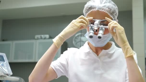 Wizyta u dentysty. Portret ortodonty przybierającego maskę i okulary powiększające lornetkę przygotowującą do zabiegu chirurgii stomatologicznej. lornetka u młodej dentystki. Nowoczesny sprzęt. 4 tys. wideo — Wideo stockowe
