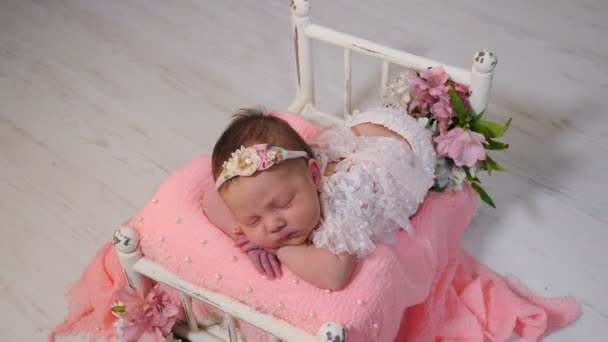 衣装を着た新生児の女の子、花で美しく装飾された小さなベッドで寝てエレガントなドレス。写真スタジオにいる赤ん坊。新生児写真の舞台裏を撮影。4 kビデオ — ストック動画