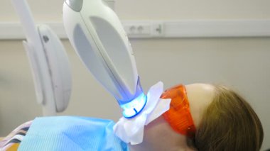 Modern diş kliniğinde profesyonel diş beyazlatma ya da beyazlatma prosedürü. LED beyazlatıcısı klinikteki sandalyede otururken koruyucu gözlüklü hasta. Ultraviyole ışınları. 4 k video