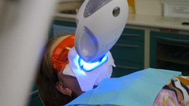 Modern diş kliniğinde profesyonel diş beyazlatma ya da beyazlatma prosedürü. LED beyazlatıcısı klinikteki sandalyede otururken koruyucu gözlüklü hasta. Ultraviyole ışınları. 4 k video