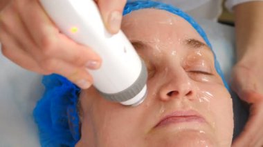 Yüz gerdirme, buruşukluk düzleştirme ve kafiye düzeltme prosedürü. Donanım kozmetoloji tedavisi. Güzellik uzmanı, kadın yüzünde ışık olan bir manipülatörü hareket ettiriyor. Cerrahi olmayan yüz güzelliği kaldırıyor.