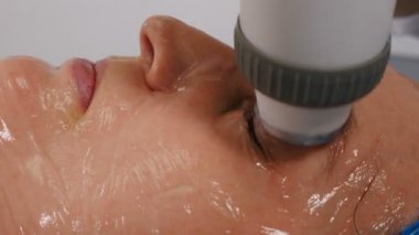 Yüz gerdirme, buruşukluk düzleştirme ve kafiye düzeltme prosedürü. Donanım kozmetoloji tedavisi. Güzellik uzmanı, kadın yüzünde ışık olan bir manipülatörü hareket ettiriyor. Cerrahi olmayan yüz güzelliği kaldırıyor.