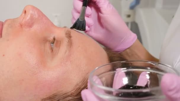 Cosmetologie, cosmetische ingrepen voor mannen. Schoonheidsspecialiste brengt donker gezichtsmasker met borstel aan. man liggend op de bank en het krijgen van gezichtshuid procedure, in schoonheidskliniek, dermatologie behandeling. 4 k video — Stockvideo