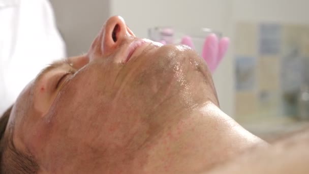Kosmetologia, zabiegi kosmetyczne dla mężczyzn. Kosmetyk używa ciemnej maski do twarzy ze szczotką. Mężczyzna leżący na kanapie i mający zabieg pielęgnacji twarzy, w klinice kosmetycznej, dermatologii. 4 tys. wideo — Wideo stockowe