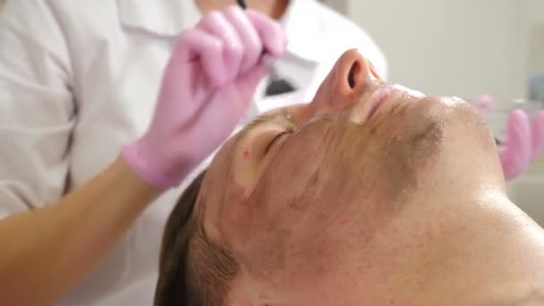 Kosmetologia, zabiegi kosmetyczne dla mężczyzn. Kosmetyk używa ciemnej maski do twarzy ze szczotką. Mężczyzna leżący na kanapie i mający zabieg pielęgnacji twarzy, w klinice kosmetycznej, dermatologii. 4 tys. wideo — Wideo stockowe