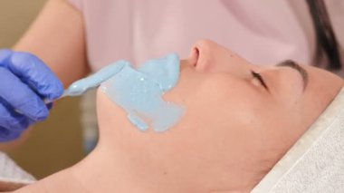 Kozmetik uzmanı aljinat maskesini dişiye spatula ile takıyor. Yüz derisi ve yaşlanma karşıtı tedavi. Kozmetoloji ve profesyonel yüz bakımı konsepti. Yüzünde aljinat maskesi olan genç bir kadın, 4 bin.