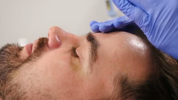 Clínica de belleza Facelifting and Anti-aging cosmetology. Hombre barbudo guapo en el procedimiento de limpieza facial realizado por cosmetóloga femenina, vista lateral. hombre recibiendo el cuidado de la piel facial rejuvenecedor. 4 k — Vídeos de Stock